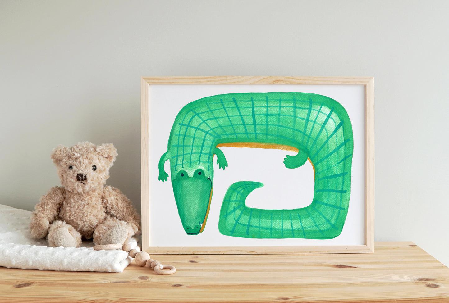 A green crocodile illustration in a wooden frame next to a plush teddy bear.