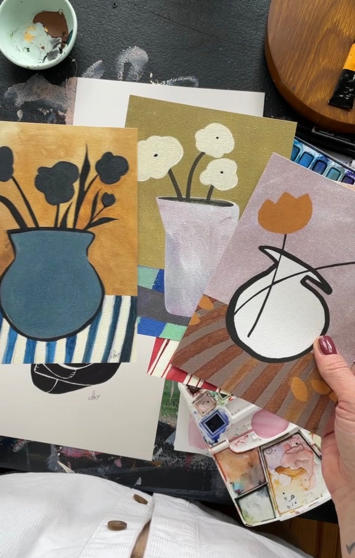 A person holding three colorful flower vase art pieces over a workspace with various art supplies.