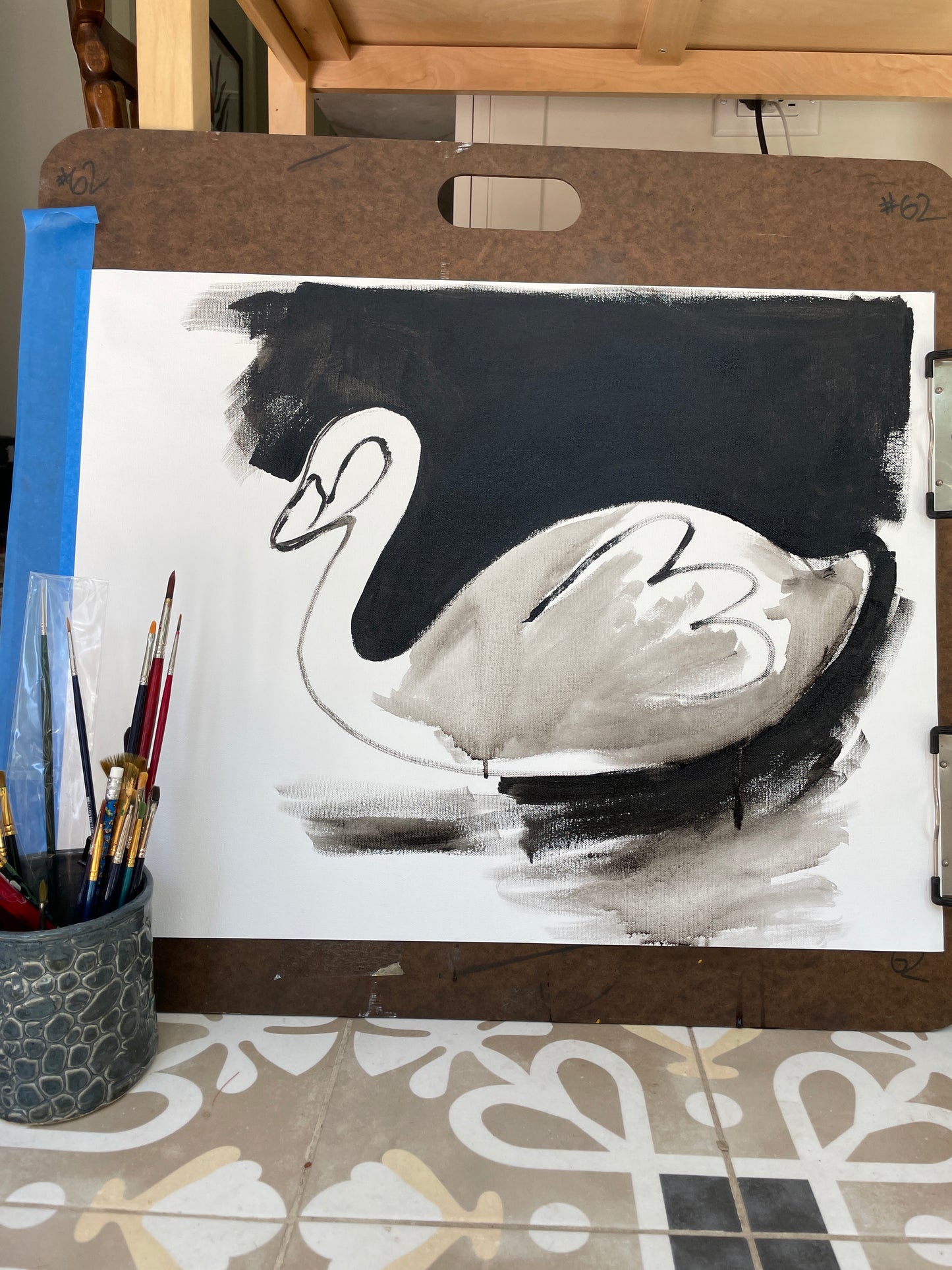 A monochromatic painting of a swan on a drawing board with paintbrushes and a pen holder in the foreground.