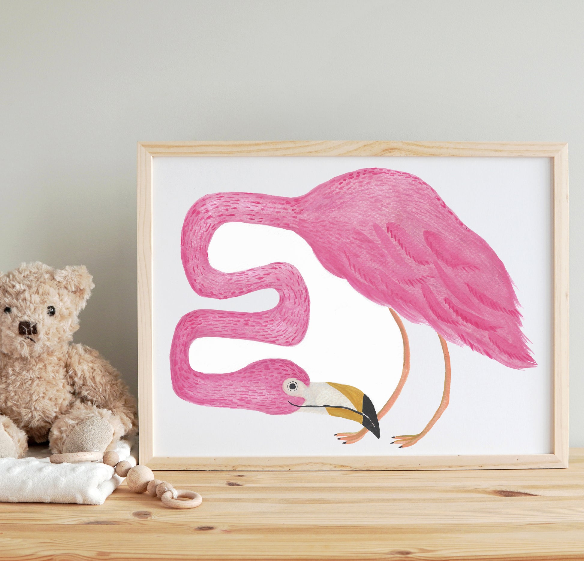 A whimsical watercolor illustration of a pink flamingo in a framed picture, displayed on a wooden table next to a teddy bear.