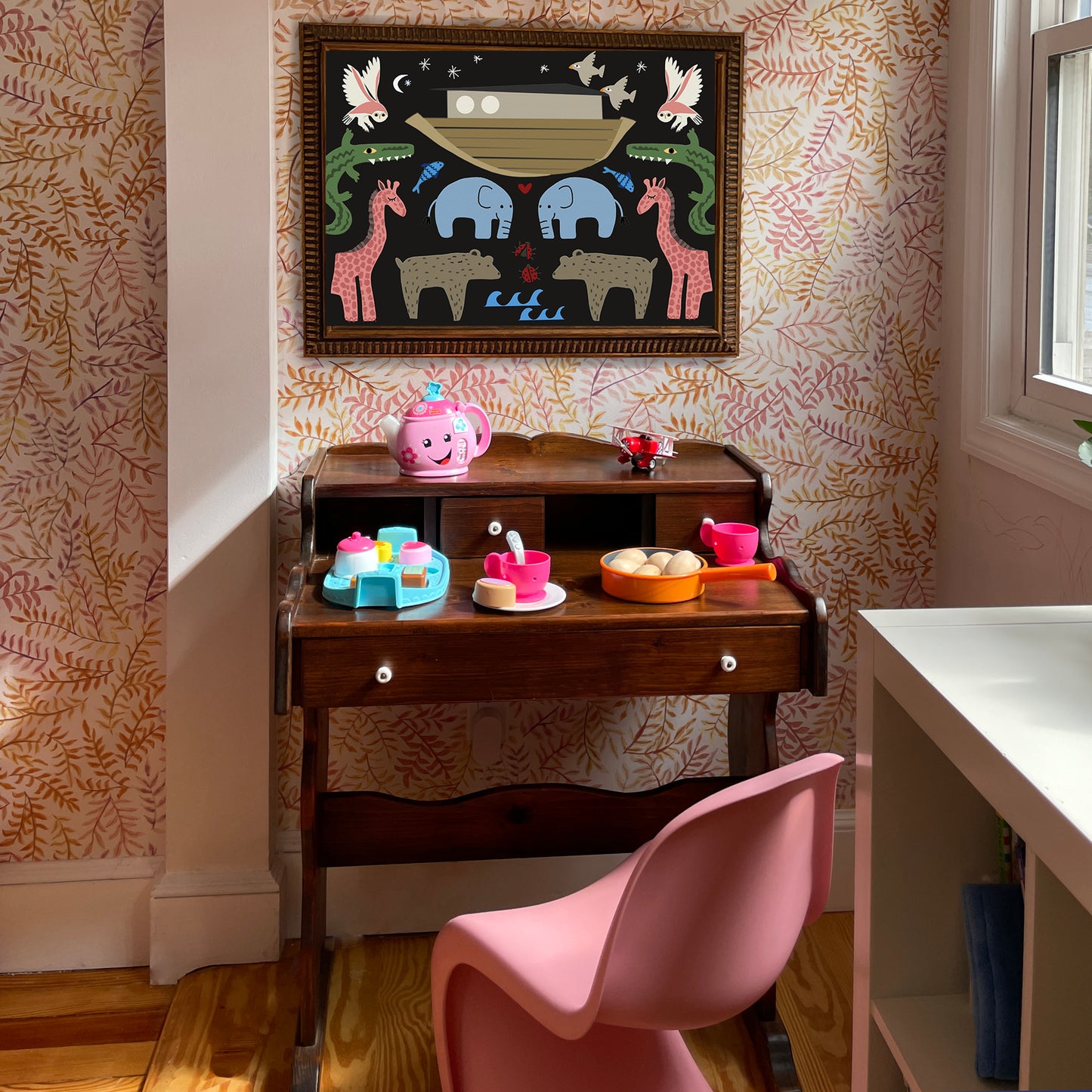 A colorful children's play area featuring a wooden desk with toys, a pink chair, and a whimsical animal-themed painting on the wall.