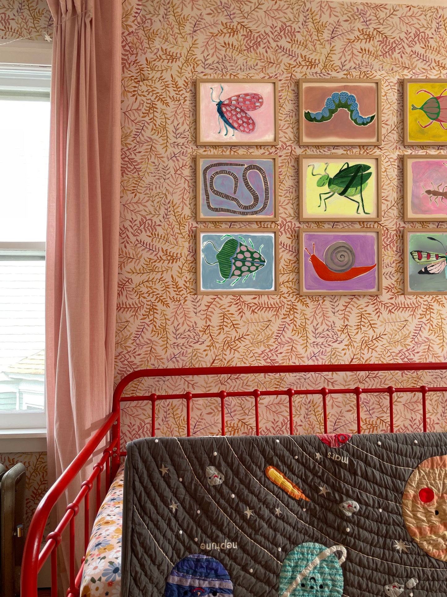 A colorful nursery featuring a red crib, a space-themed quilt, and framed artworks of insects and creatures on the wall.