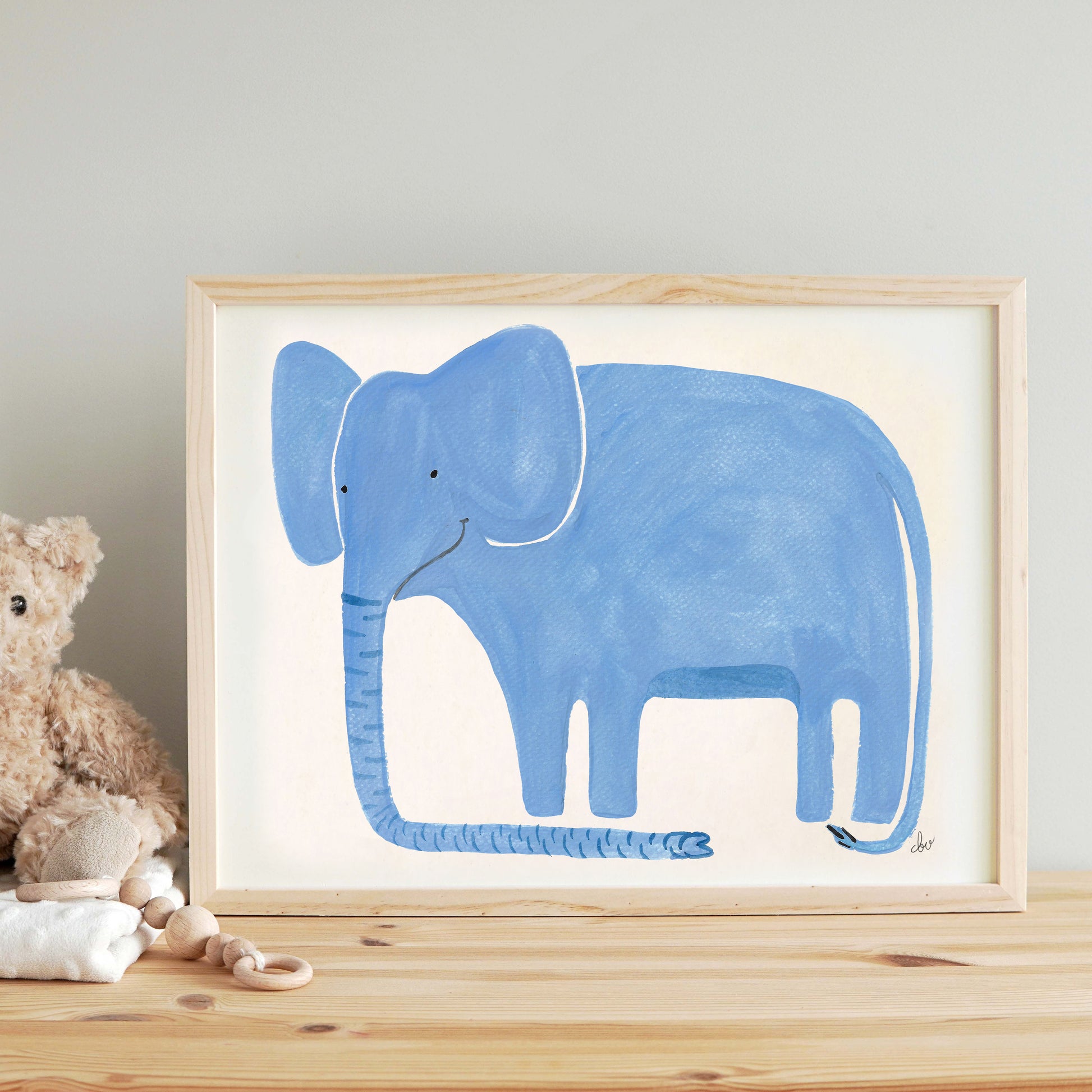 A framed painting of a smiling blue elephant on a wooden table with a plush toy and baby items nearby.