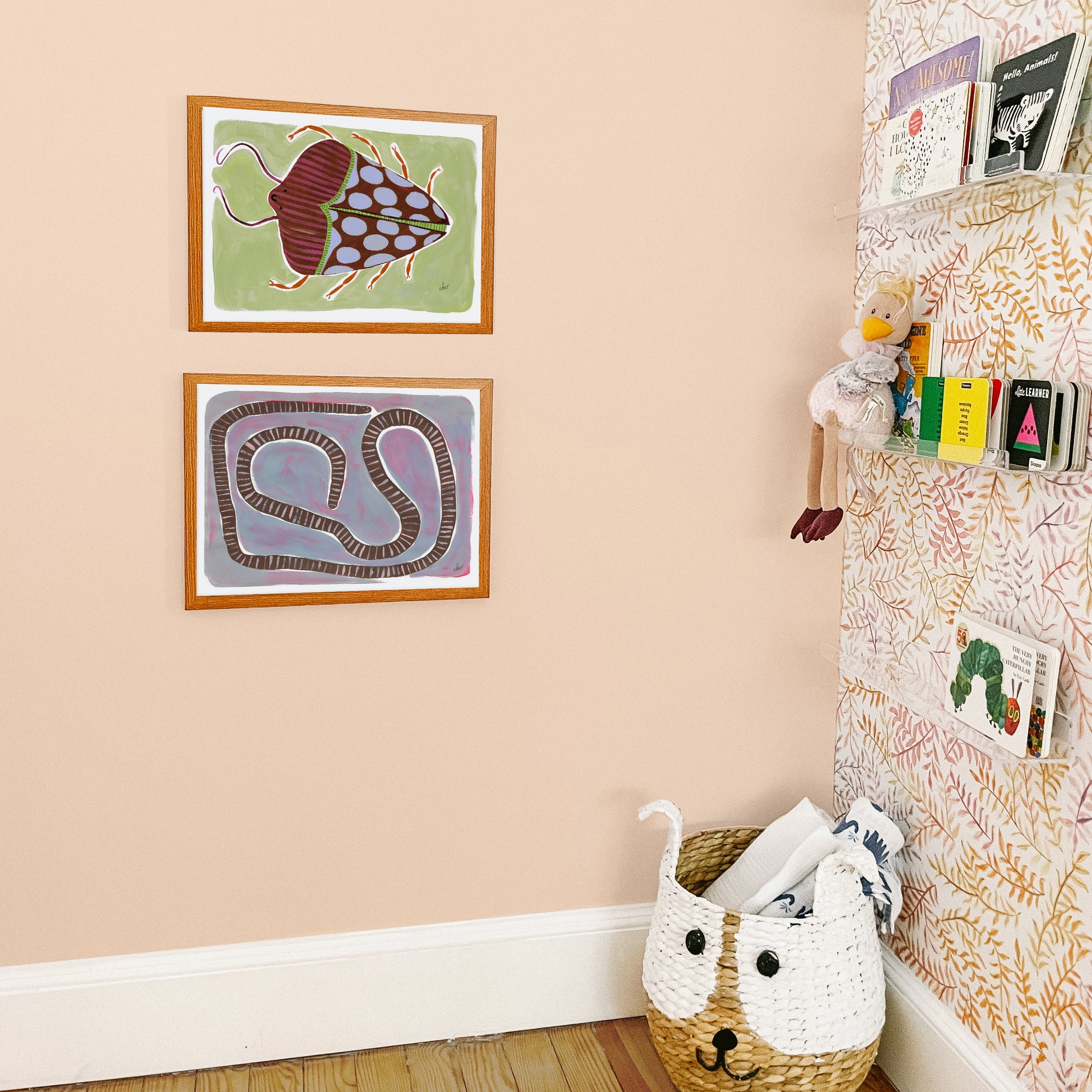 Two framed artworks featuring a fish and a snake on a peach wall, with a woven basket and wall display of small items nearby.