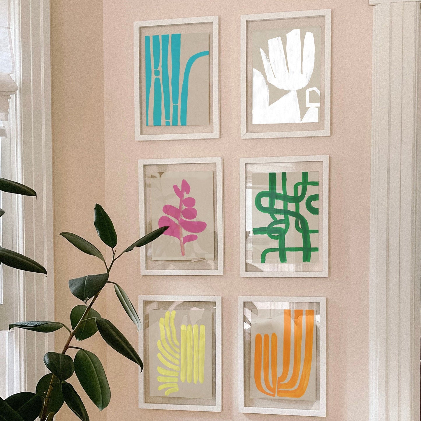 A display of six framed artworks featuring vibrant abstract designs in various colors, displayed on a light pink wall next to a green plant.