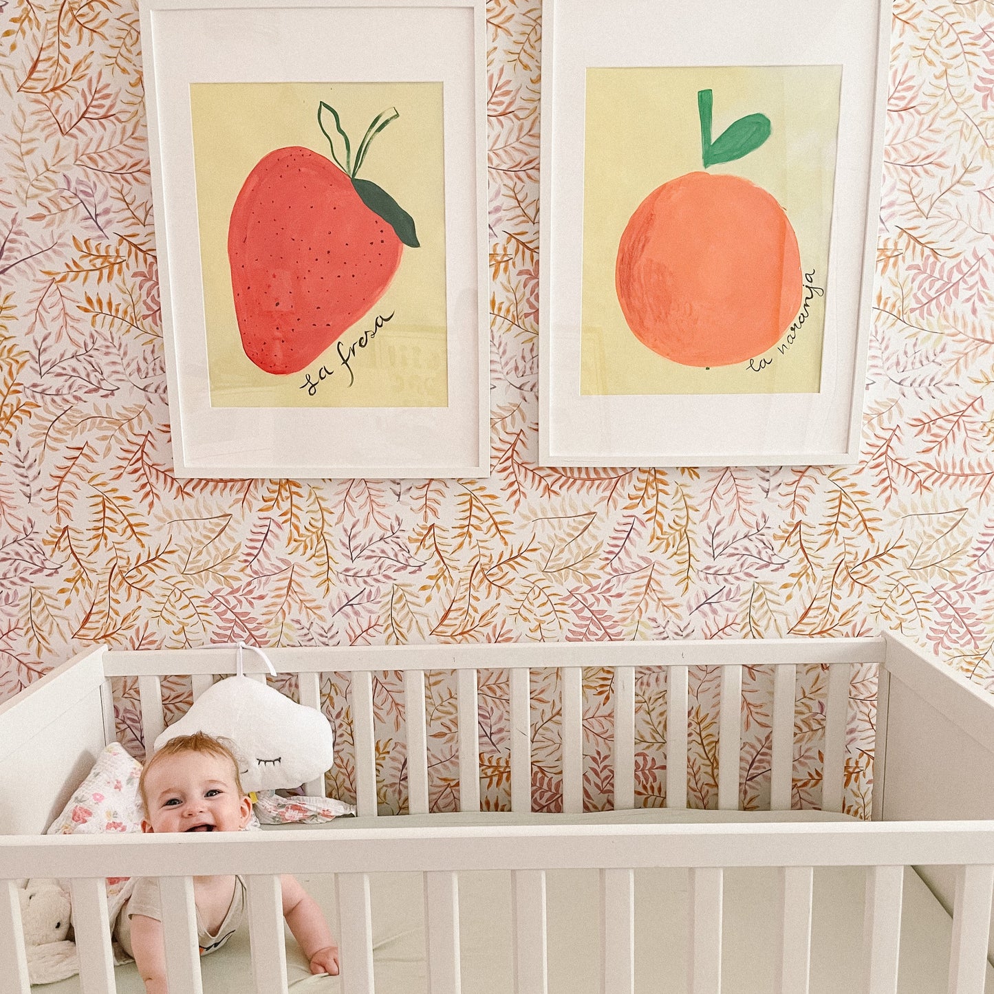 A baby in a crib with fruit-themed wall art in the background.