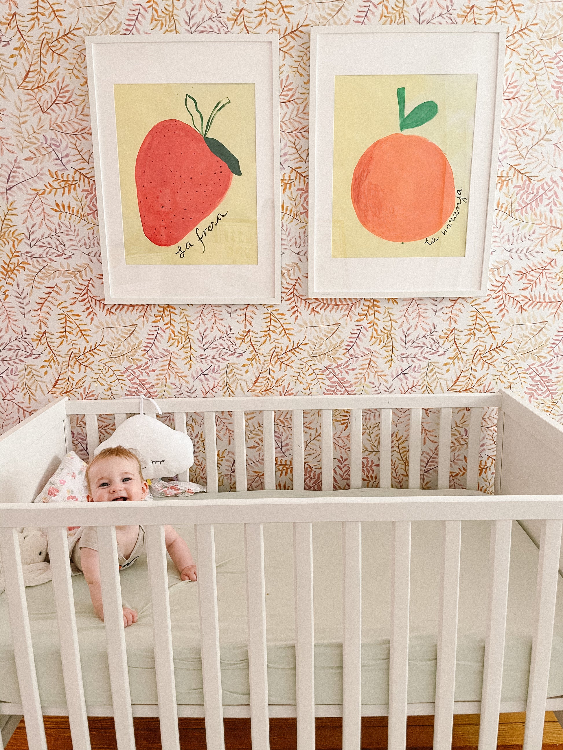 A smiling baby sitting in a white crib with colorful fruit paintings on the wall behind.