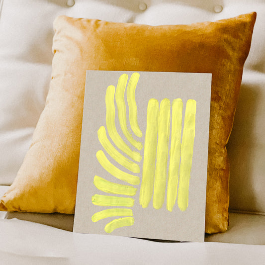 Abstract artwork with bright yellow strokes on a grey background, placed against a plush orange pillow.