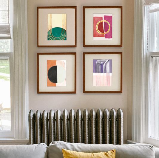 Four framed abstract art pieces displayed on a wall above a radiator, with a couch and yellow cushion in the foreground.