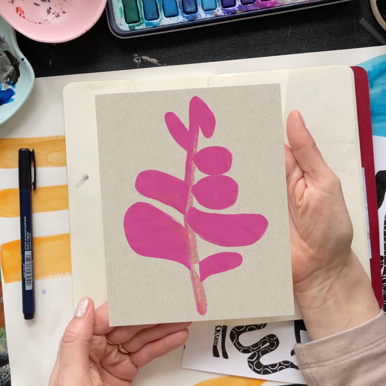 A person holding a pink leaf illustration on textured paper against an artistic workspace backdrop.